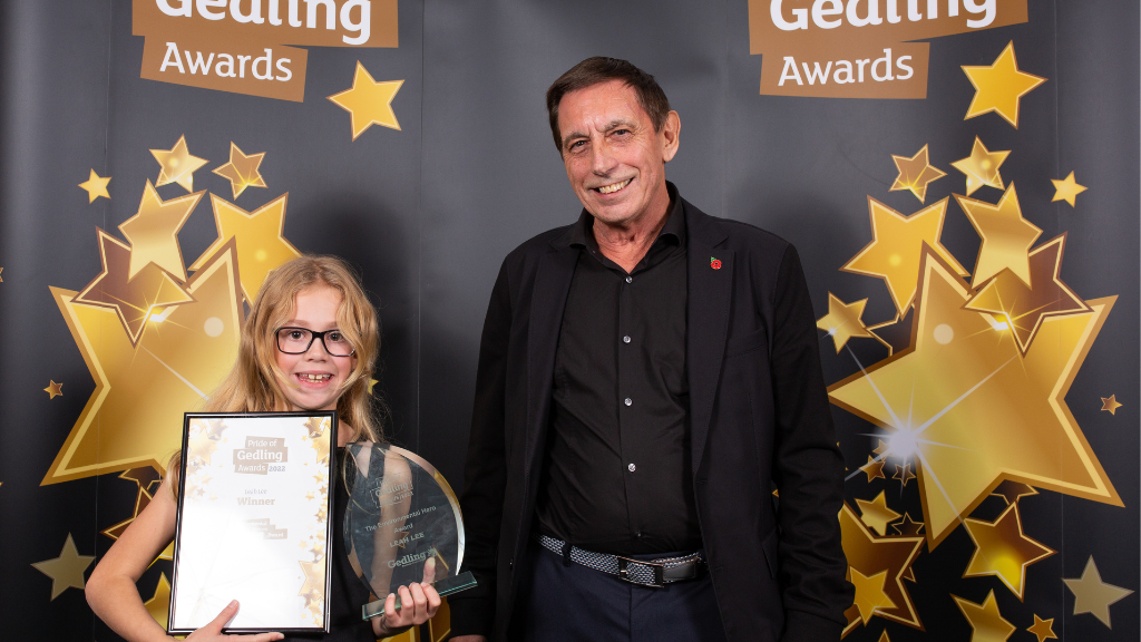 Environmental Hero winner, Leah Lee (left) holding her Pride of Gedling award and certificate in front of a black and gold Pride of Gedling banner. To her right is a representative from The Wilkins Group