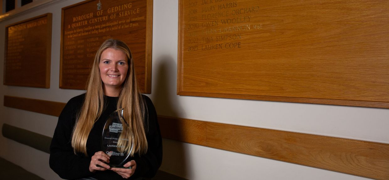 Overall Pride of Gedling Winner, Lauren Cope, holding her 2023 Pride of Gedling trophy in front of the winners board at Gedling Borough Council Civic Centre. The Board is made of wood with gold leaf names of winners written on it.